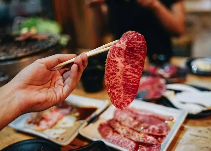 Someone holding up a thin slice of wagyu beef in chopsticks. On the table, more slices of meat.